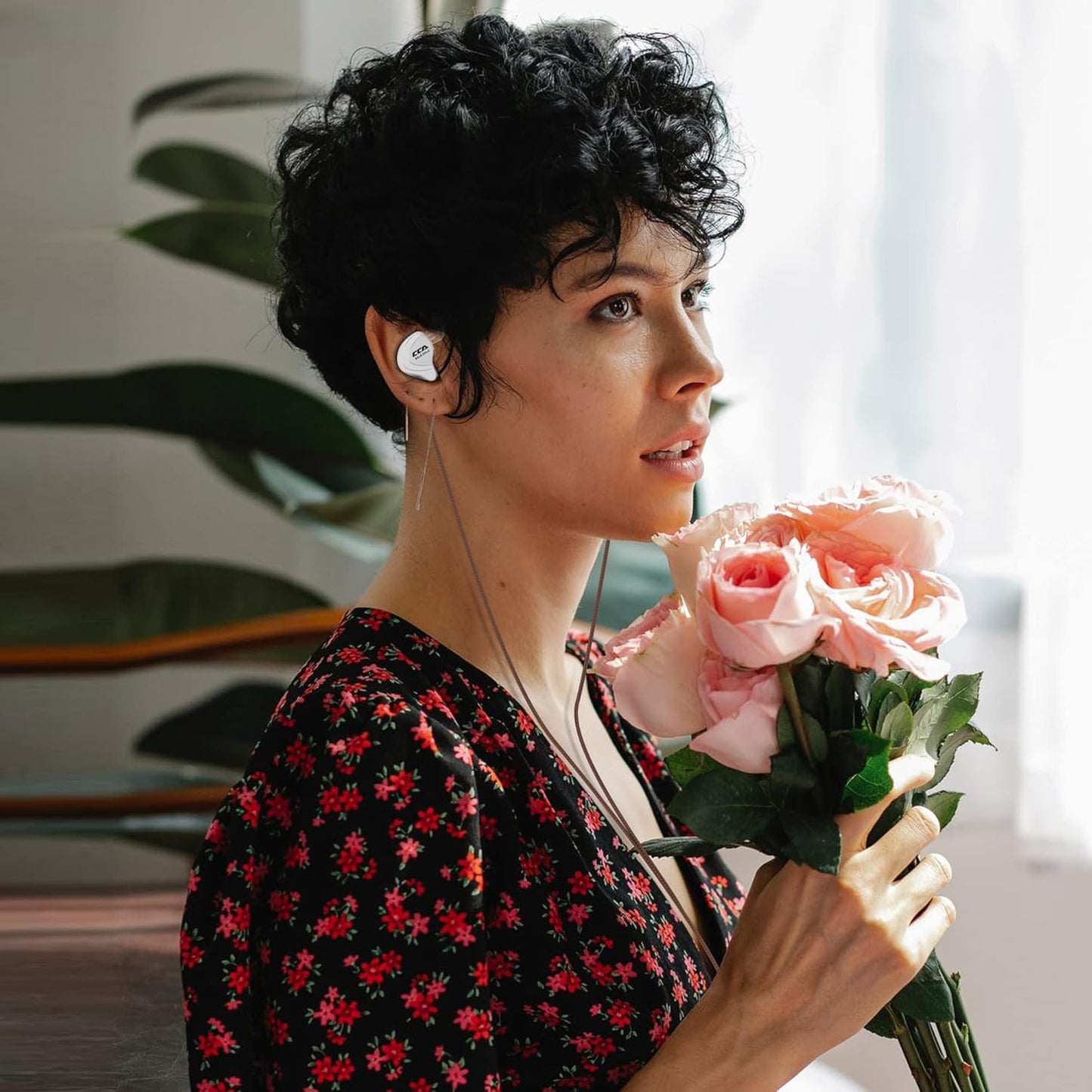 Woman holding pink roses while wearing THE VOLTRAY VAULT CCA C10 earphones, set against a bright, plant-filled background.