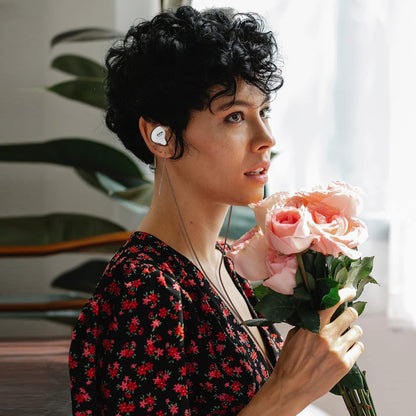 Woman holding pink roses while wearing THE VOLTRAY VAULT CCA C10 earphones, set against a bright, plant-filled background.